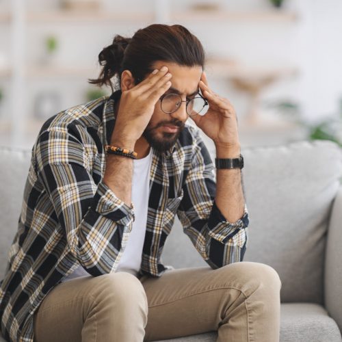 Bearded indian guy in casual with glasses sitting on couch alone at home, touching his head and looking down, suffering from headache or experiencing difficulties with job, copy space